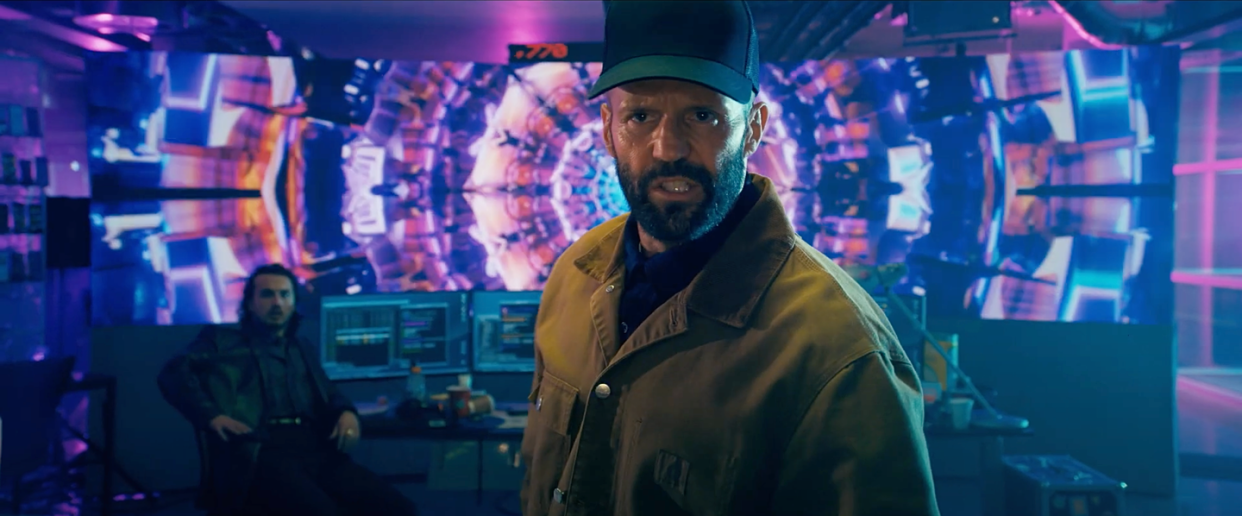 a bearded jason statham in the beekeeper, wearing a black baseball cap and a dark khaki jacket, snarling at the camera