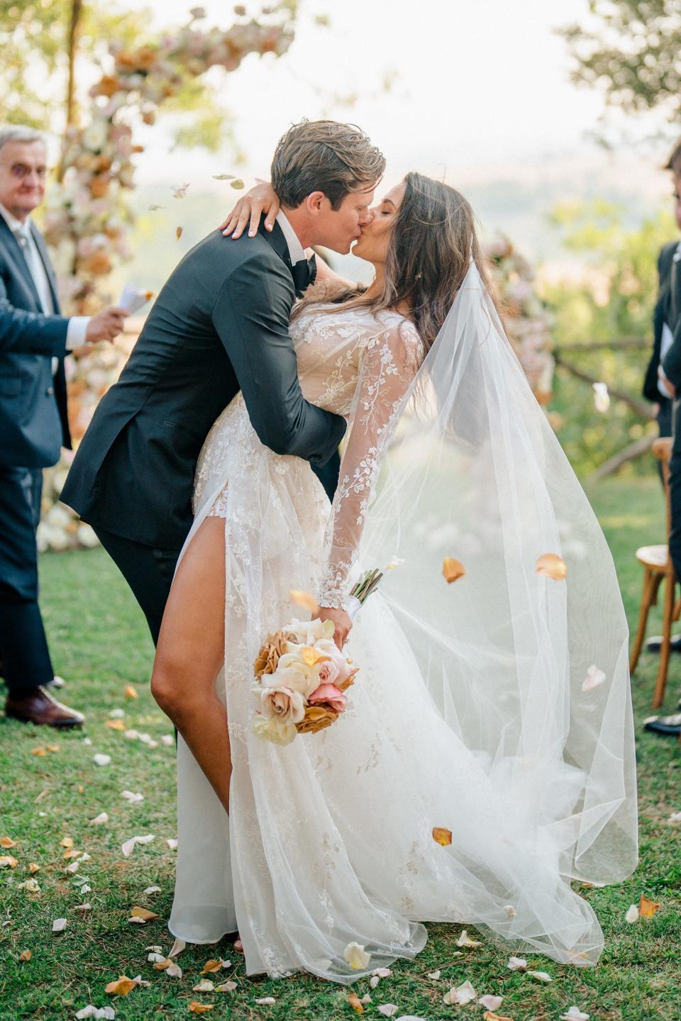 A bride and groom kiss as flower petals fly around them.