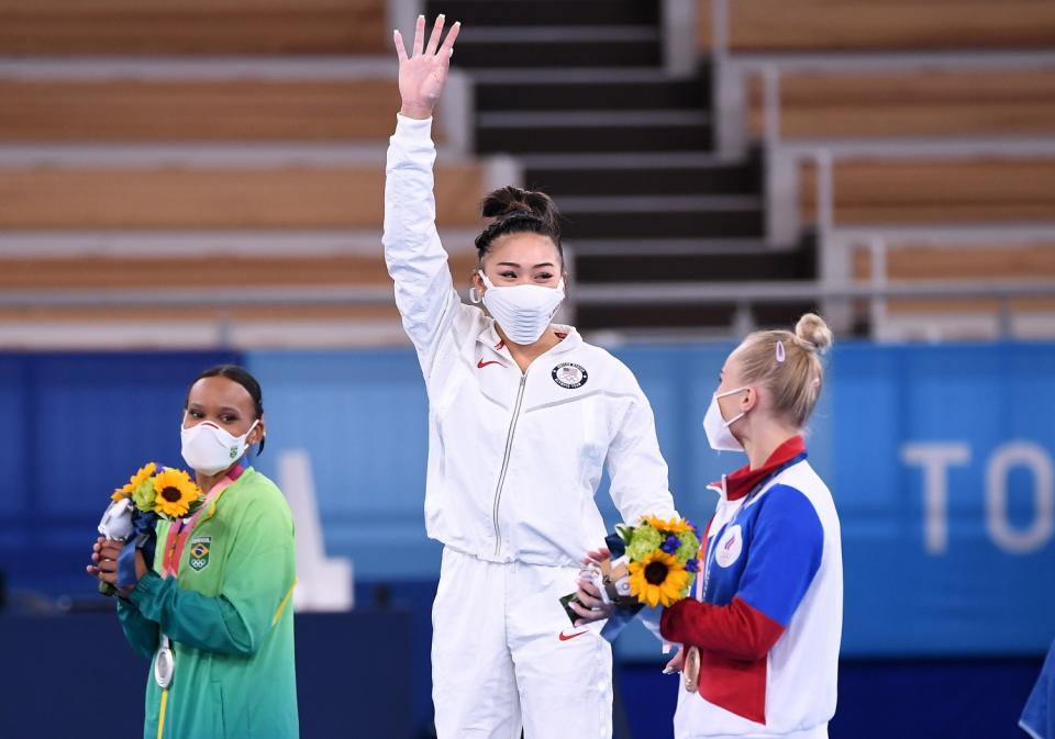 Suni Lee waves from the top of the medals podium.