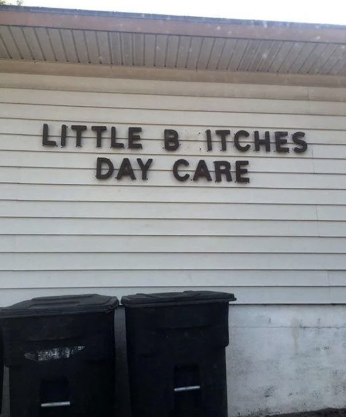 A daycare named "Little Bitches Day Care" has a sign on a building, with two trash bins placed below it