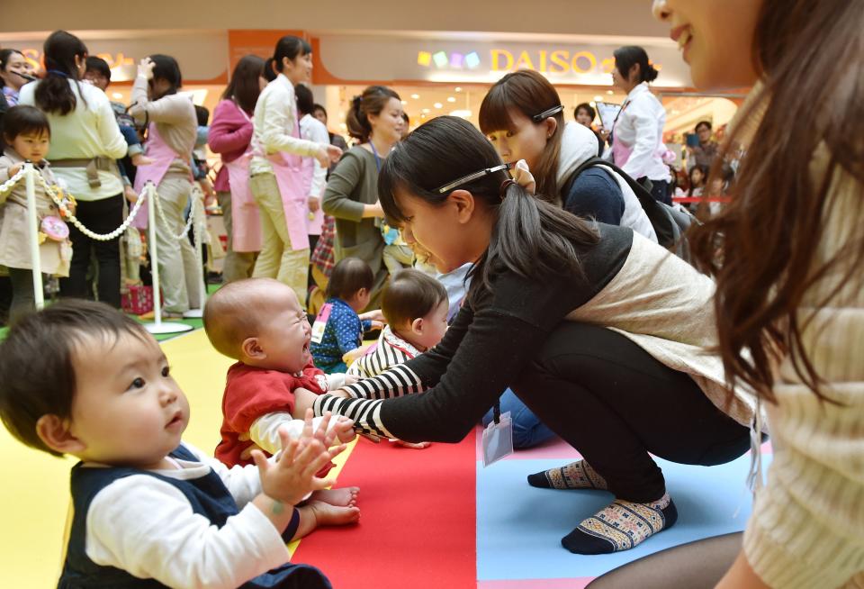 A group of women interacting with babies who are sitting on colored mats at the start of a baby-crawling competition.