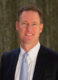 Salesforce President and COO Brian Millham is shown smiling to the camera and wearing a navy suit, white shirt and light blue tie.