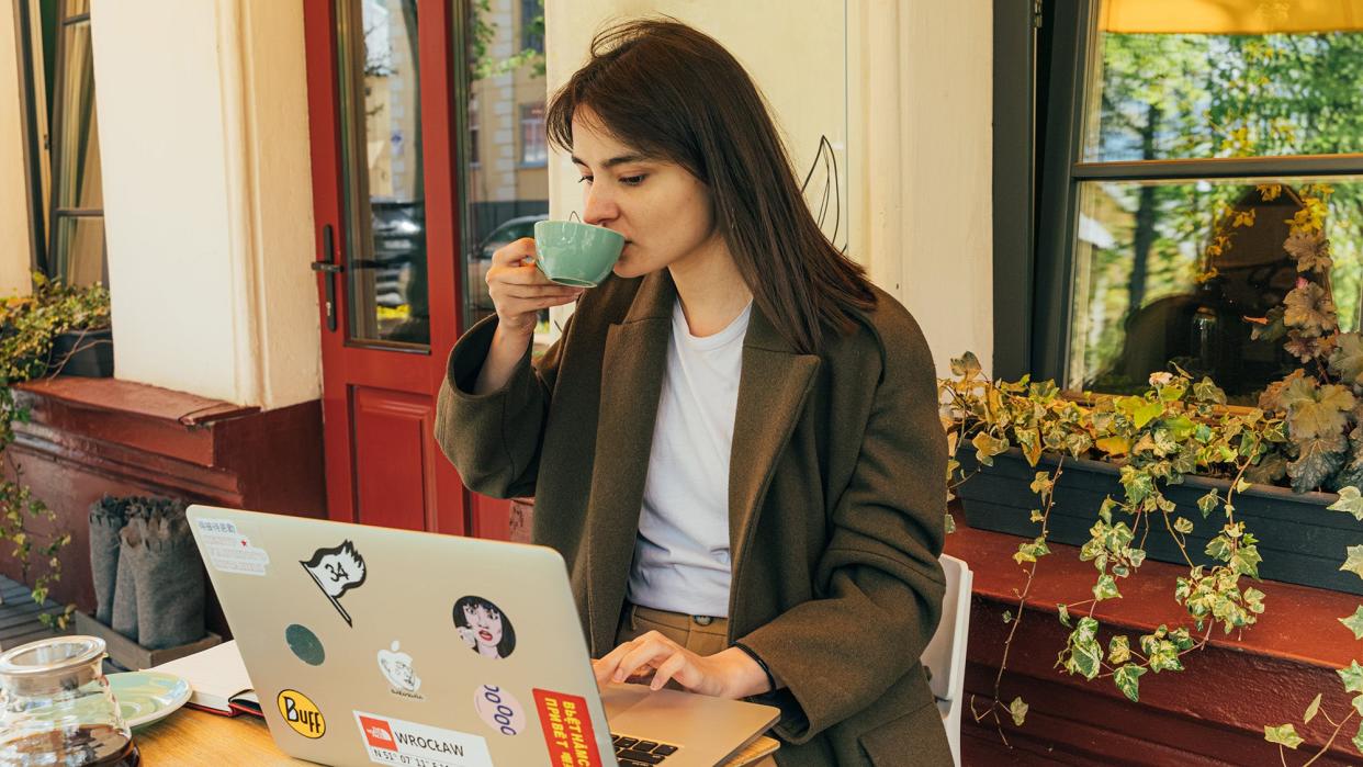 Person sitting outside of a coffee house on a sunny day, drinking from a green cup and reading from an open MacBook.