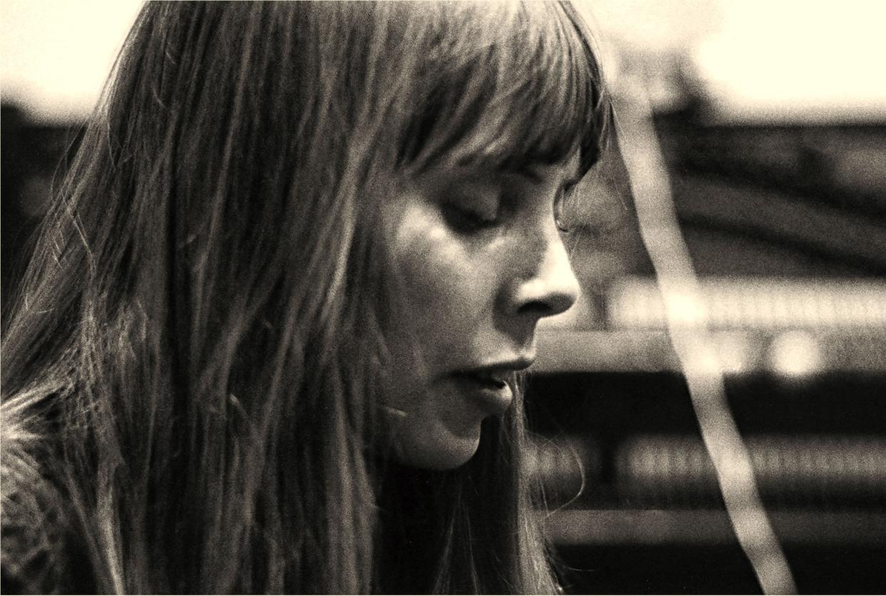 Joni Mitchell singing and looking down at the keyboard of a grand piano she is playing in the recording booth, profile, with an upright piano in the background.