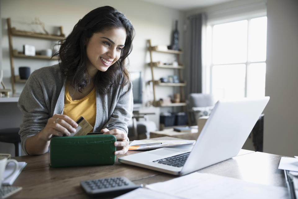 Smiling young woman with credit card and laptop paying bills online in dining room