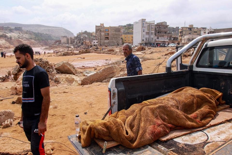 Two men walk past the body of a flash flood victim, which is covered by a blanket and lying in the back of a pickup truck, in Derna.