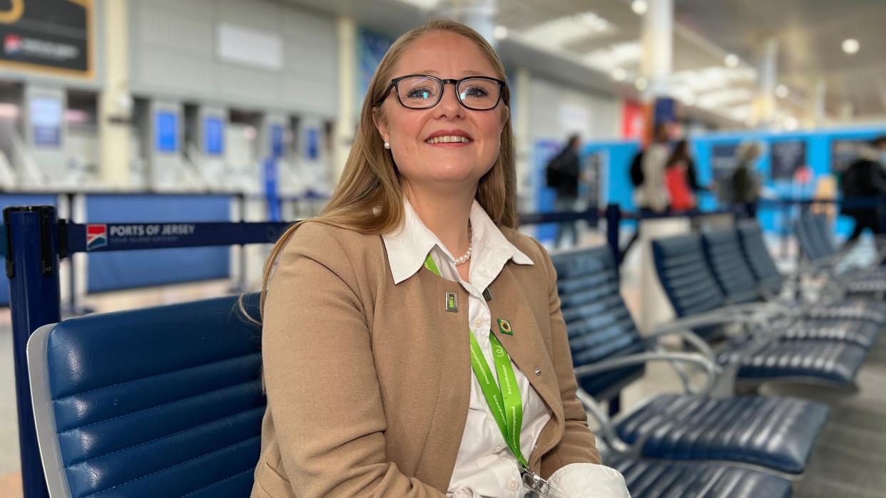 Sarah-Louise Stubbs smiles at the camera as she sits on a blue chair and in the background people are heading the depatures section of the airport