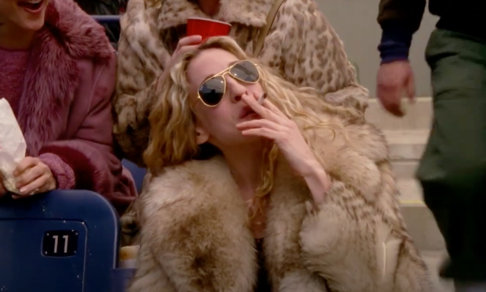 Woman in fur coat and sunglasses smoking while seated, holding a red cup. Two other people are partially visible next to her