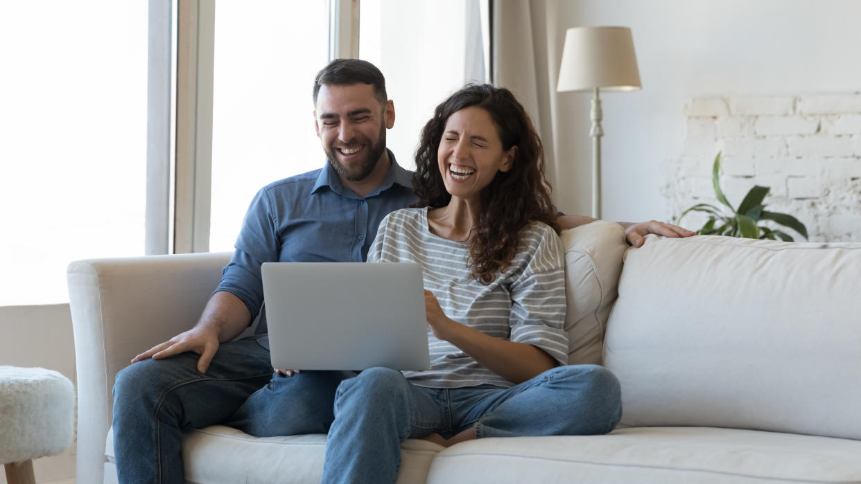 Happy overjoyed millennial couple using laptop at home together, resting on comfortable sofa, laughing, feeling joy, receiving good news, talking on video call. Communication concept