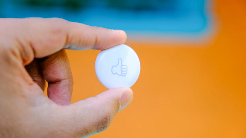 A person’s left hand holding an Apple AirTag with a thumbs-up emoji laser-engraved onto it.