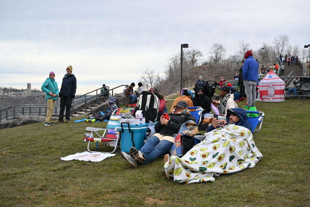 A dozen people on a low hill in winter clothes, some on lawn chairs.