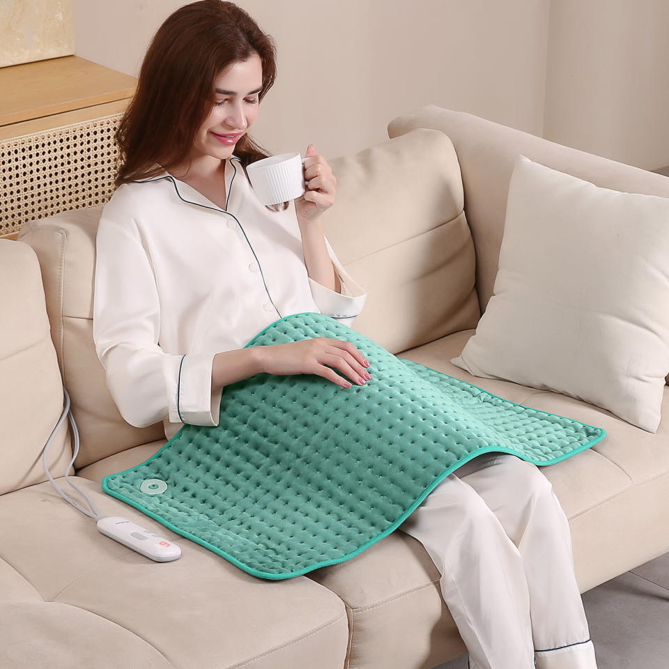 a woman on the couch drinking tea with her green heating pad