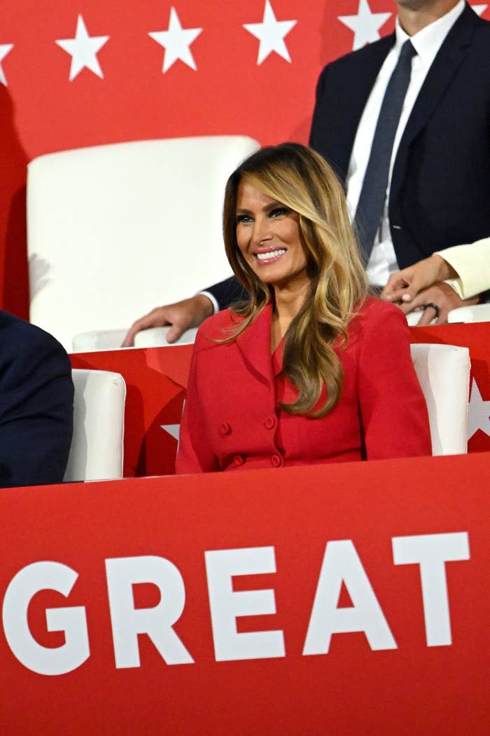 Melania Trump in a red outfit smiles and sits in front of a red background with white stars. The word "GREAT" is partially visible