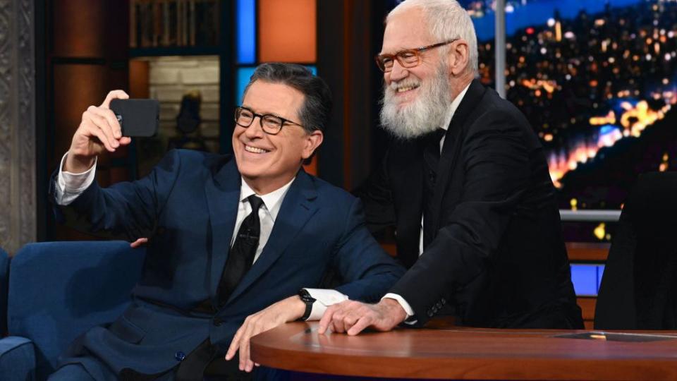 stephen colbert sitting down and holding his phone out for a selfie with david letterman