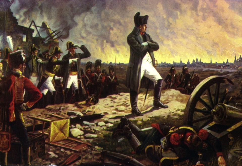 A painting of Napoleon Bonaparte standing with one foot forward, looking over the battlefield with his army behind him. At his feet are dead bodies and broken cannons.