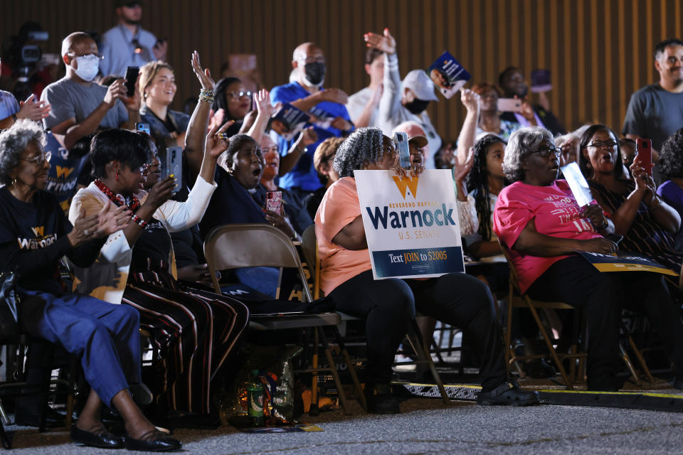 A few dozen people on folding chairs applaud and hold their hands up. One holds a sign that reads: Warnock, U.S. Senate