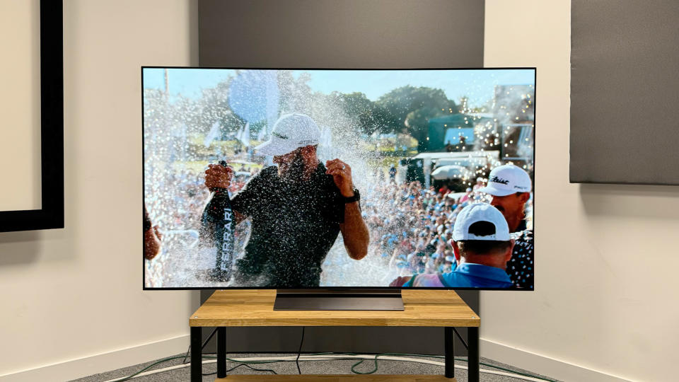 65-inch LG C4 TV photographed straight-on on a wooden stand. On the screen is an image of a golfer being sprayed with champagne.