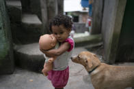 FILE - In this March 24, 2020 file photo, a girl holds her doll in an alley of the Rocinha slum of Rio de Janeiro, Brazil, amid orders to stay home to contain the spread of the coronavirus. In Rocinha, narrow alleyways reduce airflow around homes packed tightly together; clean water is scarce, sewage often runs in the streets and winding alleys and soaring staircases make it difficult for medical professionals to retrieve an ailing patient in case of emergency. (AP Photo/Leo Correa, File)