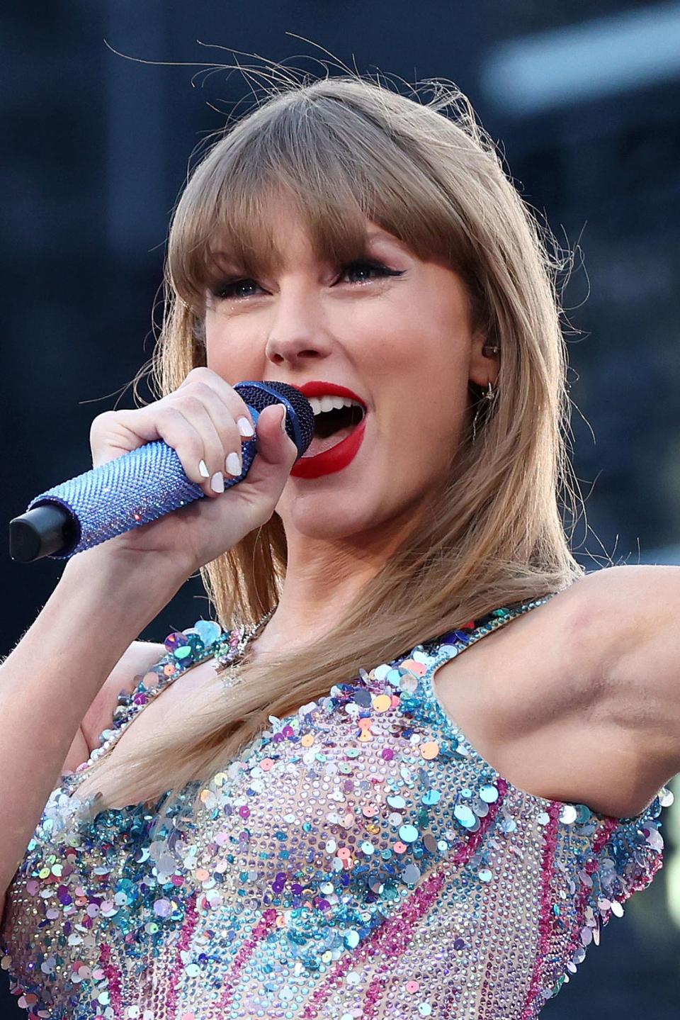 Taylor Swift performing on stage, wearing a sparkling outfit, singing into a microphone, hand raised