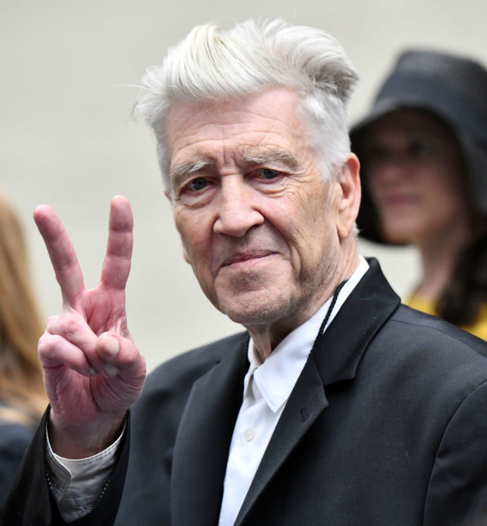 David Lynch flashes a peace sign while wearing a black jacket over a white shirt. A woman with her face obscured and wearing a hat is in the background