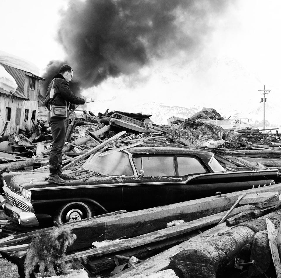 File - In this March 29, 1964 file photo, a photographer looks over wreckage as smoke rises in the background from burning oil storage tanks at Valdez, Alaska, March 29, 1964. The city was hit hard by the earthquake that demolished some of Alaska's most picturesque and largest cities. North America's largest earthquake rattled Alaska 50 years ago, killing 15 people and creating a tsunami that killed 124 more from Alaska to California. The magnitude 9.2 quake hit at 5:30 p.m. on Good Friday, turning soil beneath parts of Anchorage into jelly and collapsing buildings that were not engineered to withstand the force of colliding continental plates.(AP Photo/File)