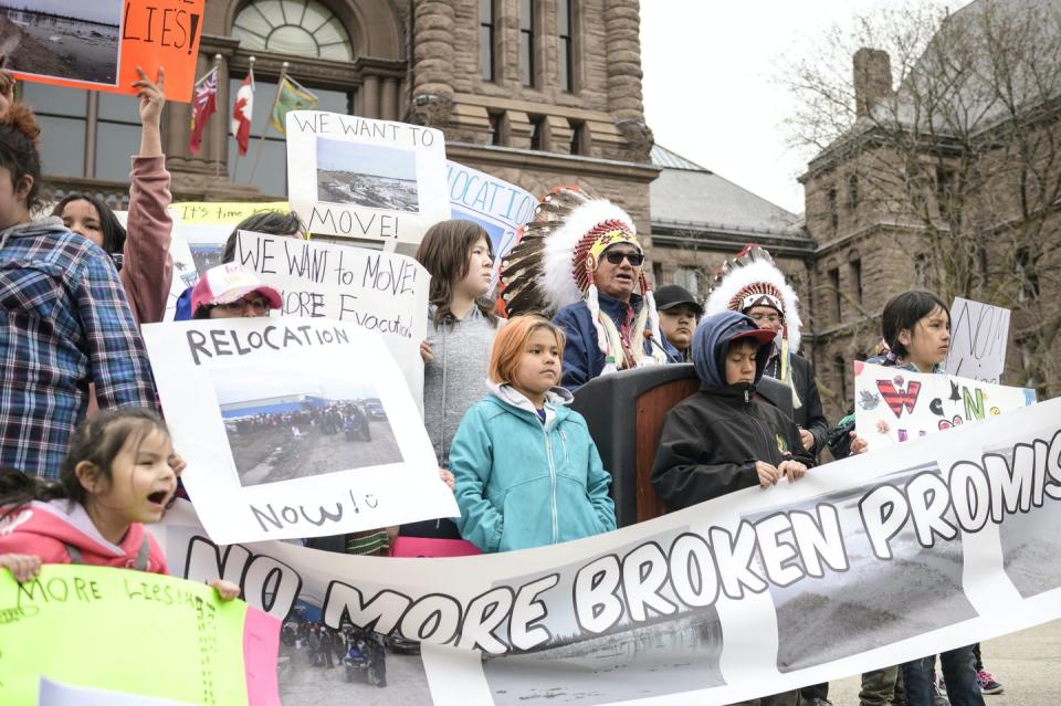 A group of people stand holding signs that read 'no more broken promises' and 'relocation now'