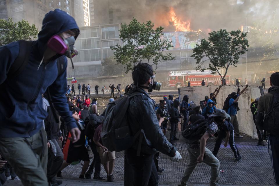 Anti-government protestors walk toward police as firefighters work to put out a fire set by protesters at a shopping center in Santiago, Chile, Monday, Oct. 28, 2019. Fresh protests and attacks on businesses erupted in Chile Monday despite President Sebastián Piñera's replacement of eight important Cabinet ministers with more centrist figures, and his attempts to assure the country that he had heard calls for greater equality and improved social services. (AP Photo/Rodrigo Abd)