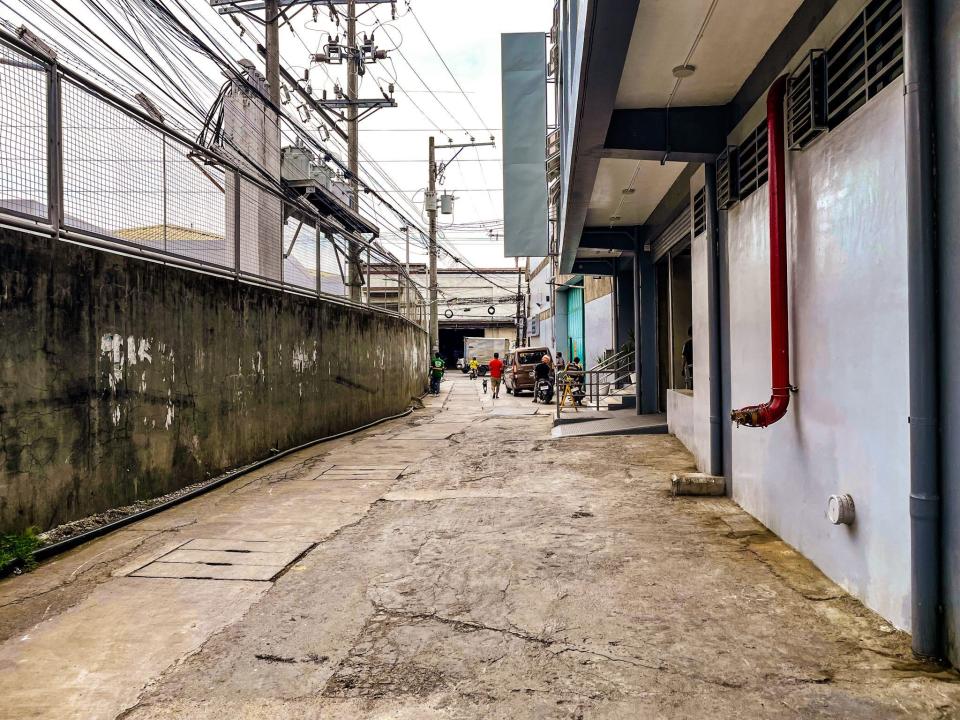 A street with a large concrete fence with telephone cables above it on one side and a line of buildings on the other.