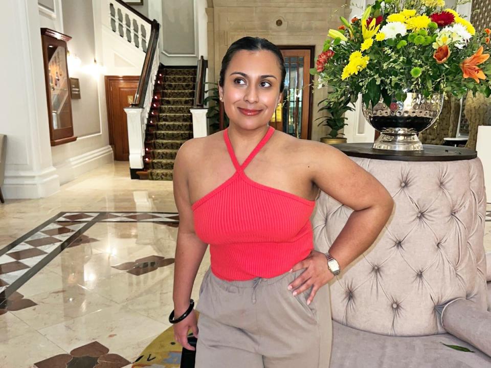 The writer wears a bright-red top and beige pants and holds a hand on her hip in a hotel lobby. The lobby has gray seating, a staircase, and a vase filled with green, yellow, and orange flowers