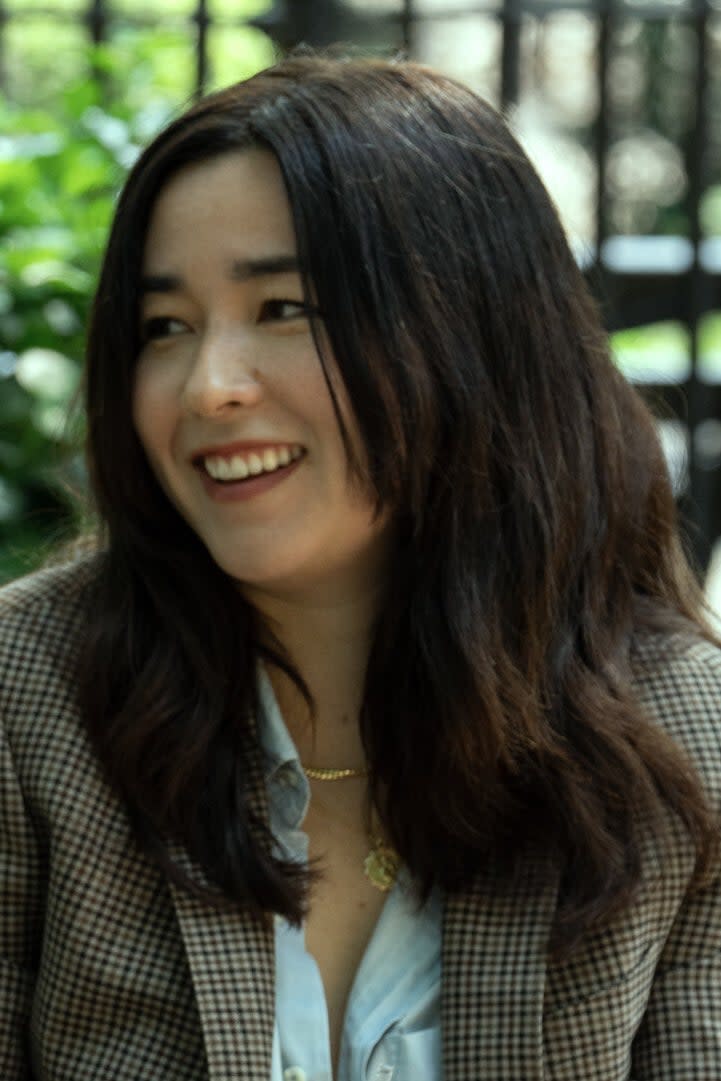 Donald Glover in a leather jacket and Maya Erskine in a blazer sit on a park bench, smiling and chatting. Glover holds a snack, while Erskine holds a drink