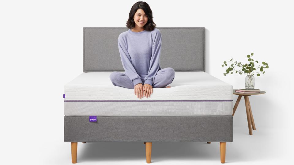 A woman with dark hair and dressed in lilac coloured pyjamas sits on the edge of the Purple Plus Mattress Purple Plus Mattress