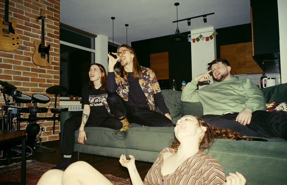 Four friends laughing and lounging casually on a green couch in a cozy living room with guitars on the wall