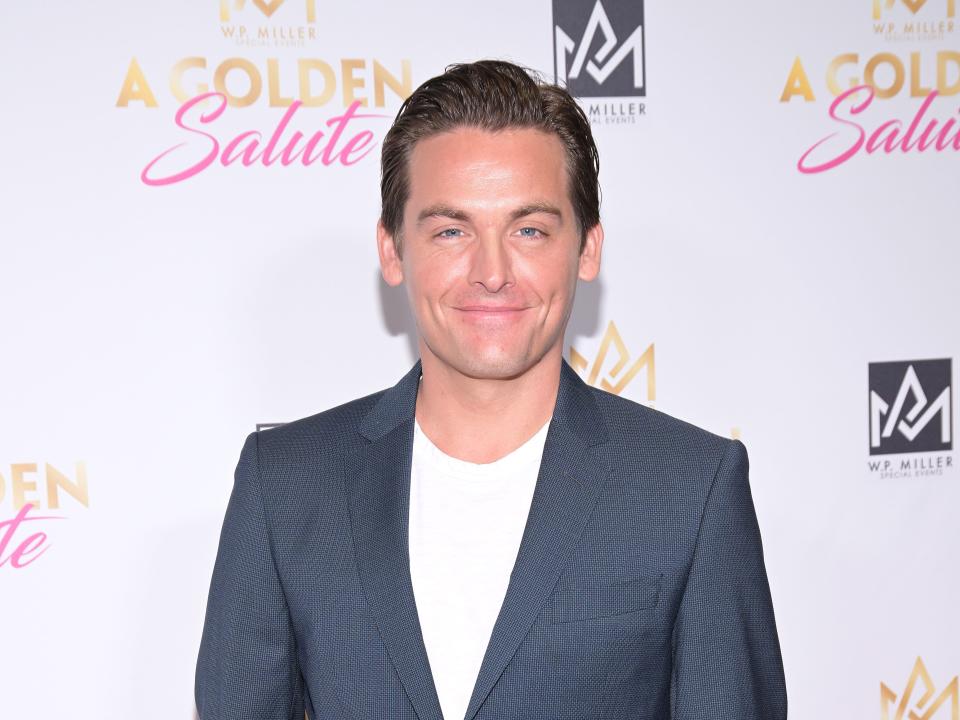 kevin zegers wearing a grey suit over a white tshirt, smiling and standing with his hands in his pockets on a red carpet