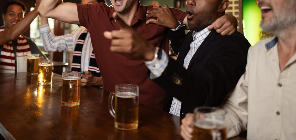 Men cheering and smiling while watching a sports game in a bar, with drinks and a soccer ball on the counter