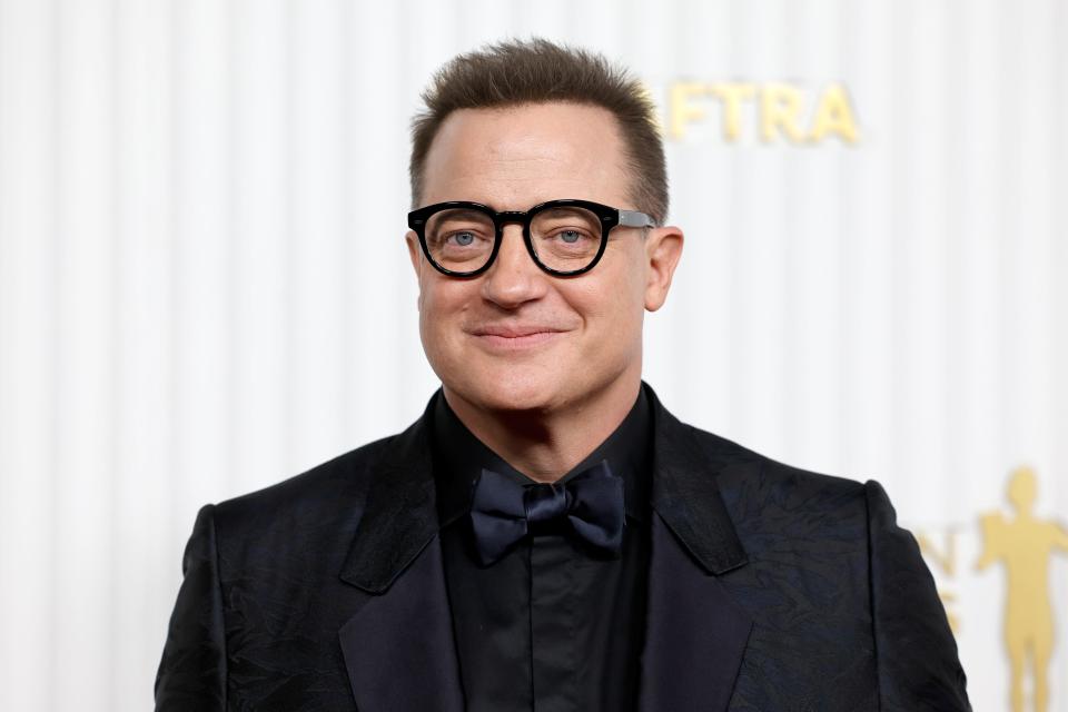 Brendan Fraser wears a black jacket, shirt, bowtie, and glasses in front of a white, striped background at the 2023 SAG Awards in Los Angeles.