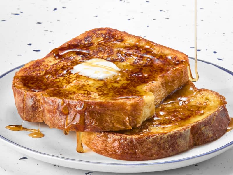 angled shot of two pieces of french toast on a white plate with a blue rim, topped with a pat of butter and drizzled with maple syrup