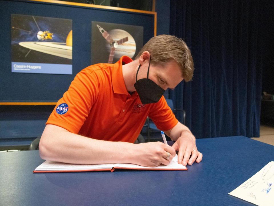 havard grip man in orange nasa shirt black kn95 sits at blue table writing in book