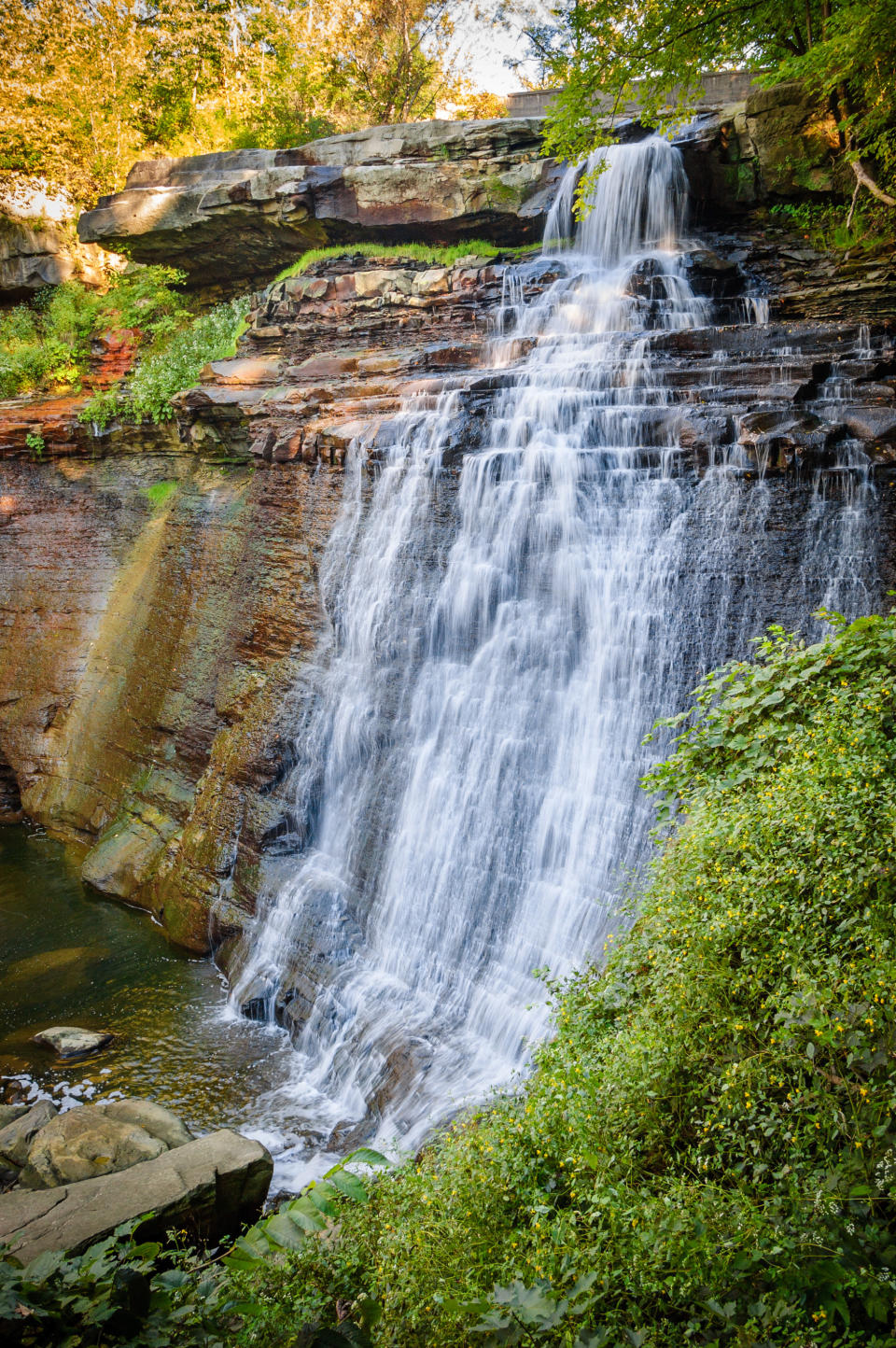 A tall waterfall rushes over a worn out rock formation, made up of flat layers of various thickness. Lush bushes and leaves surround the waterfall.