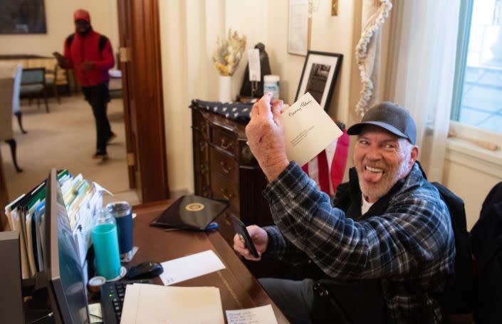 Richard Barnett sentado en un escritorio con un sobre con el nombre de Nancy Pelosi mientras saca la lengua.