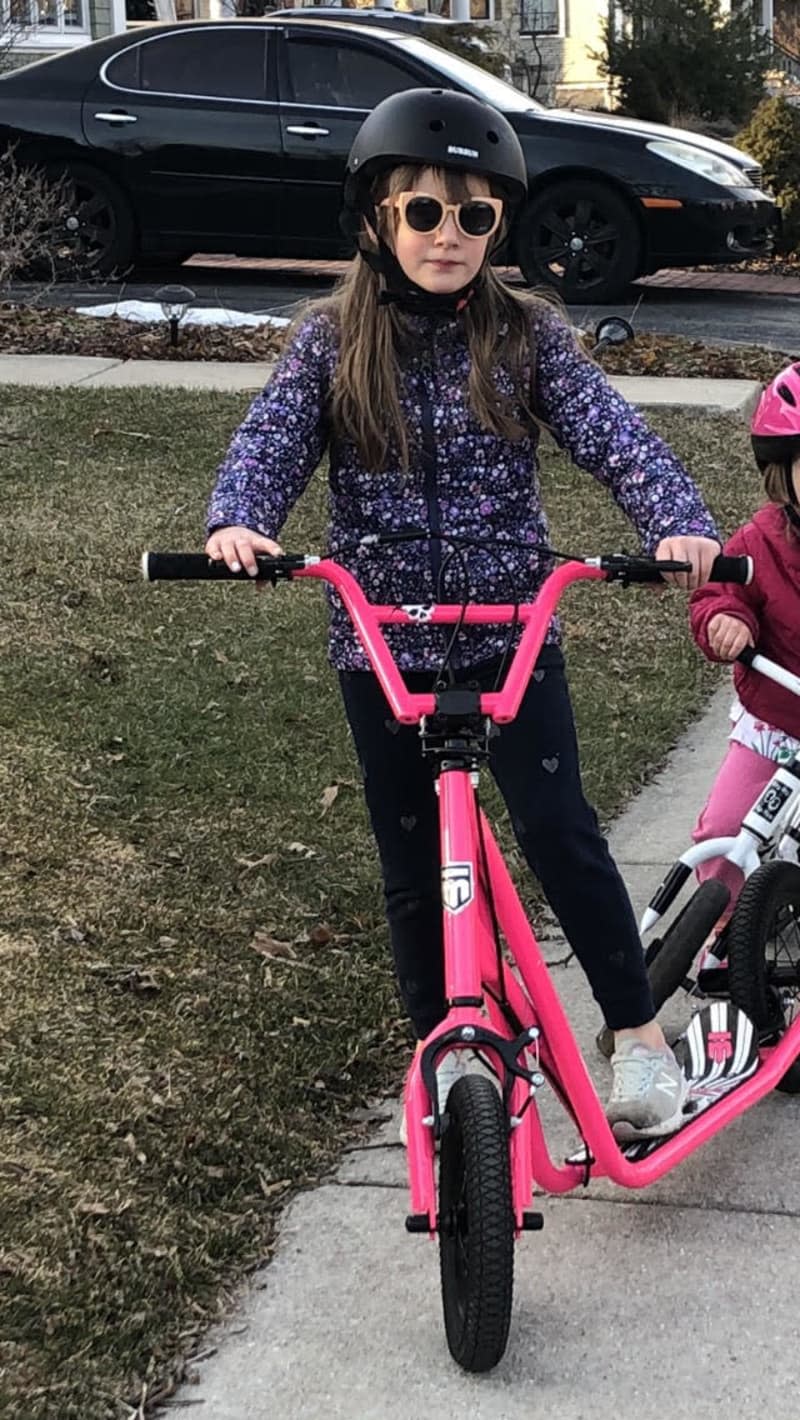 Girl in black helmet and sunglasses riding a pink Mongoose Youth Expo scooter