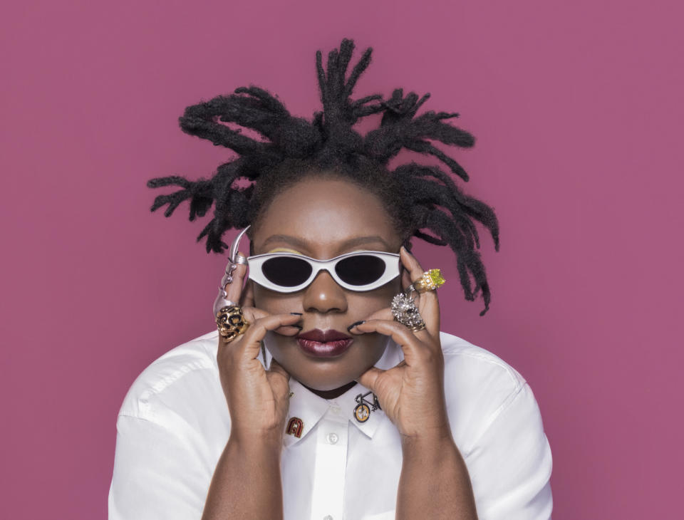 Shot of designer Mimi Plange against a pink background.  She is wearing a white colored shirt with vintage pins, white sunglasses and several rings on both hands.