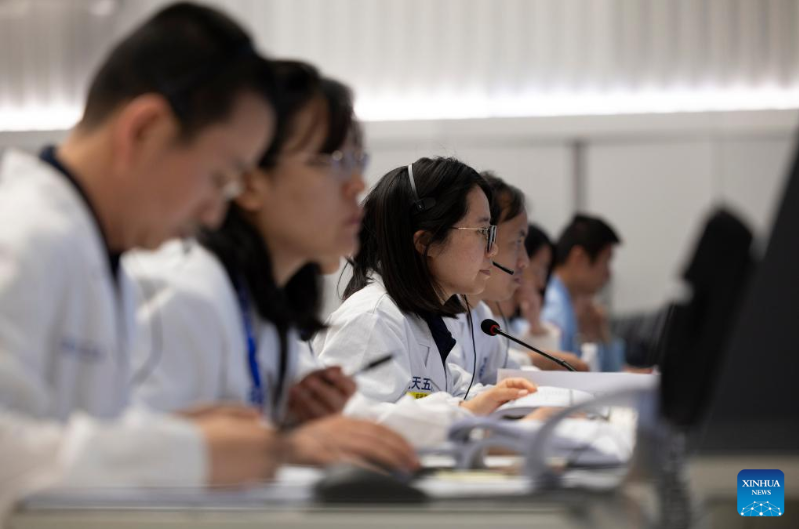 Six people in white outfits sit in a mission control room, all looking in the same direction.