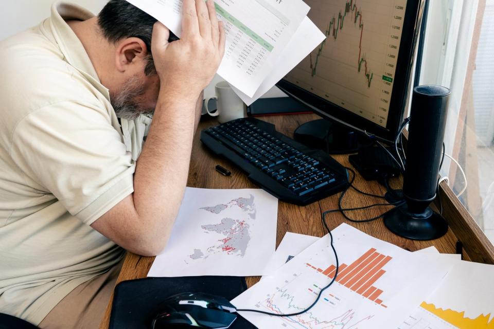 Frustrated person looking at documents on a desk that also has a computer screen showing a chart. 