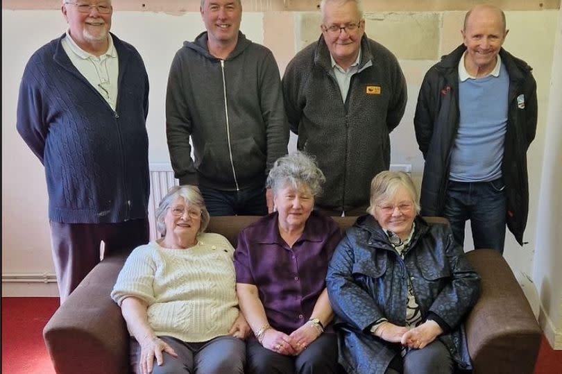 Group of people lined up with four standing behind a sofa with three sitting on it