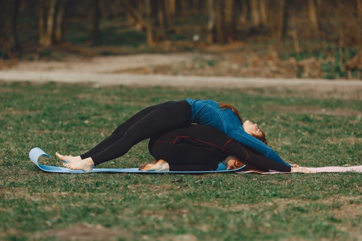 A woman in dark clothes folds forward in Child's pose while her yoga partner in a blue shirt does a back bend over her back. They are spine to spine. They are practicing outside on grass.