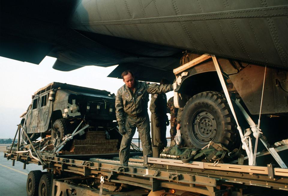 A man stands on a platform with his hand on a truck being loaded into the back of a plane