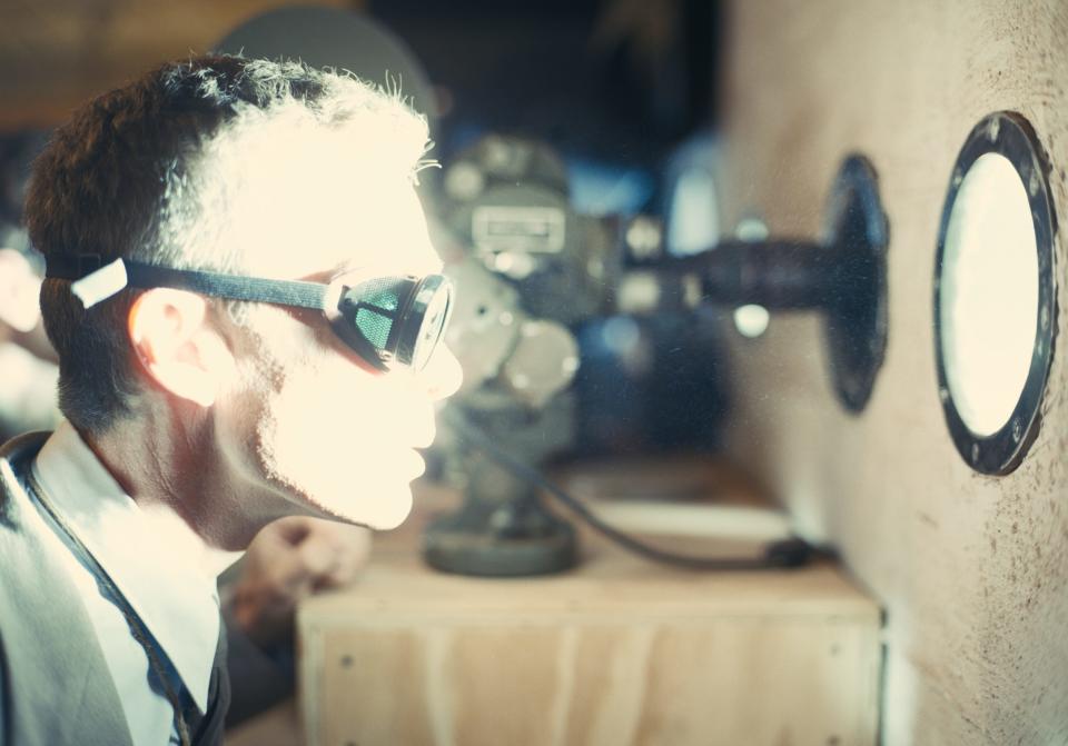 Cillian Murphy in a scene from "Oppenheimer" where Oppenheimer is seen wearing a set of googles and looking through a porthole while a bright light illuminates his face.