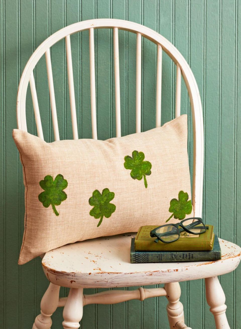 a linen pillow with green four leaf clovers painted on the front resting on a white chair behind a small stack of green books