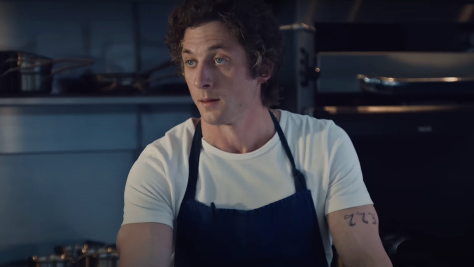 Jeremy Allen White in a  t-shirt and a dark apron stands in a kitchen, with a tattoo of 