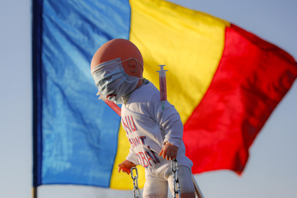 Anti-vaccination protesters hold a doll with "I don't want to be a lab rat" written on it backdropped by a Romanian national flag, during a rally outside the parliament building in Bucharest, Romania, Sunday, March 7, 2021. Some thousands of anti-vaccination protestors from across Romania converged outside the parliament building protesting against government pandemic control measures as authorities announced new restrictions amid a rise of COVID-19 infections. (AP Photo/Vadim Ghirda)